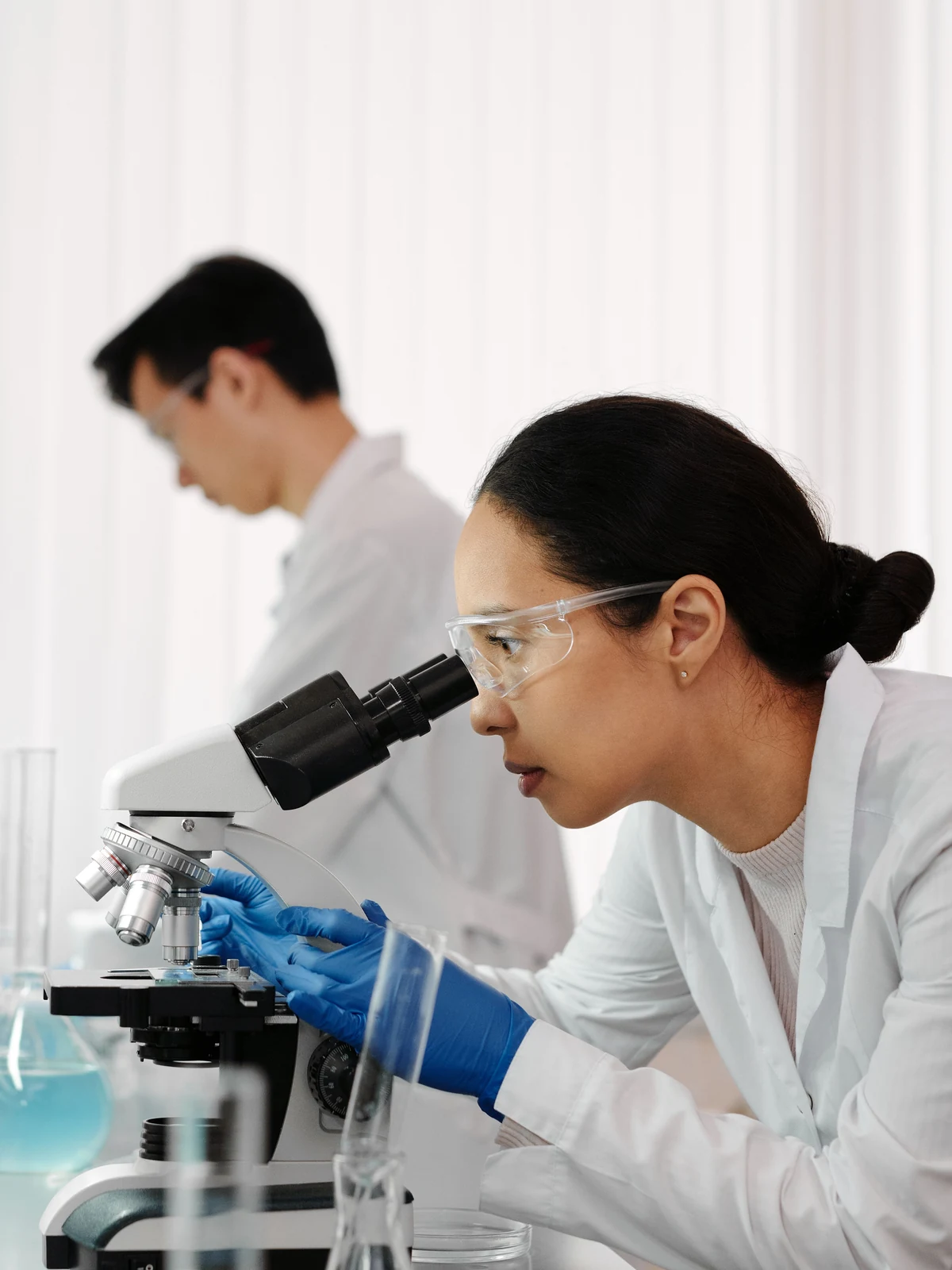 Two researchers at a laboratory bench, one examining a sample under a microscope